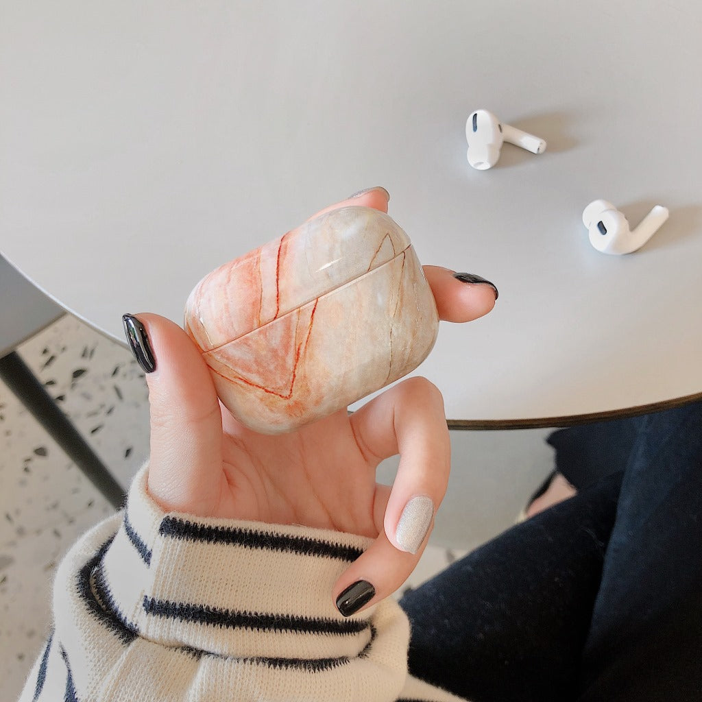 Marble-patterned airpod case held by a hand with black nail polish, with airpods on a table in the background.