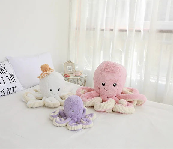 Three plush octopus toys in white, pink, and purple on a bed with a light background.