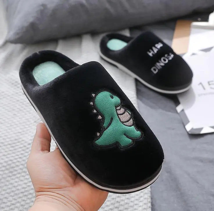 Black slippers with green dinosaur design held by a hand on a gray surface.