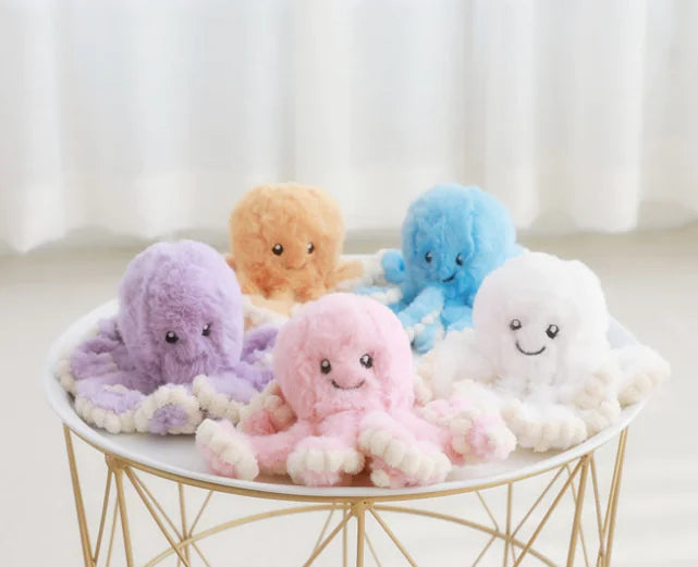 Five colorful plush octopus toys on a small round table with a white curtain background.
