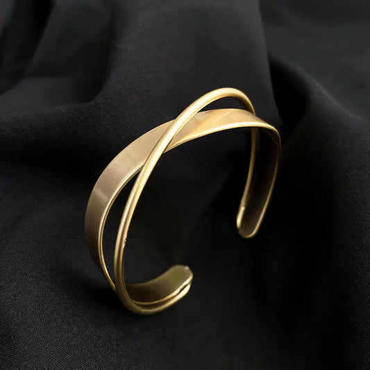 Gold-plated wave-shaped bangle with cross detail resting on a neutral fabric backdrop.