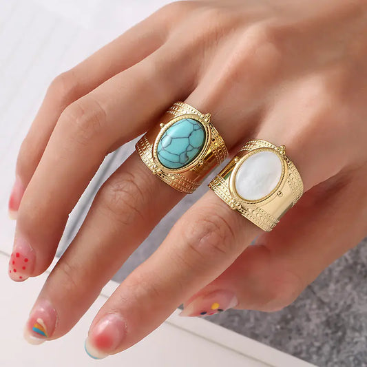 Hand wearing two gold rings with turquoise and white stones on a neutral background